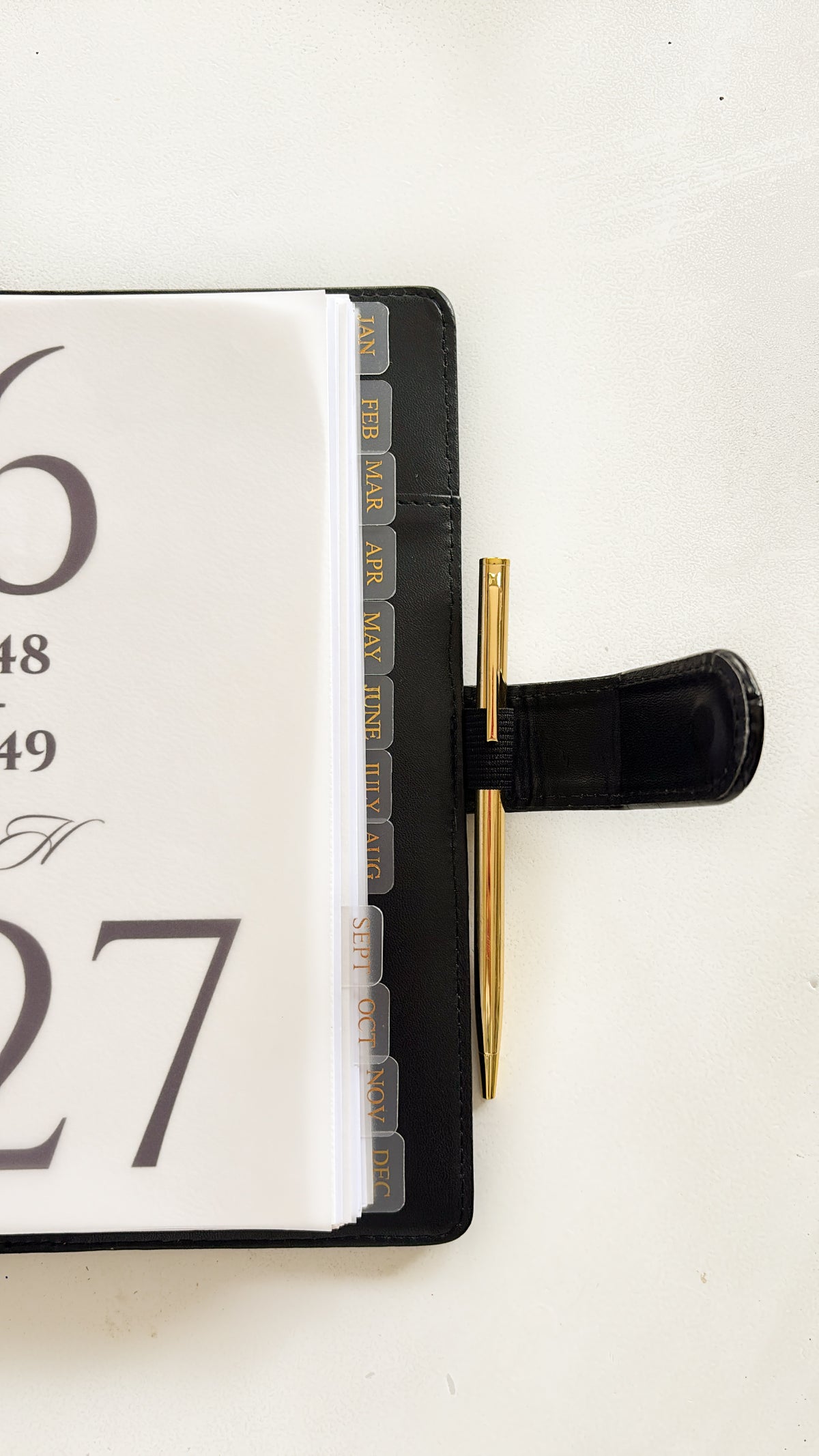 A close-up of a black leather planner or desk organizer with a calendar page inserted and a gold pen tucked into a black loop on the right side, set against a plain off-white background.