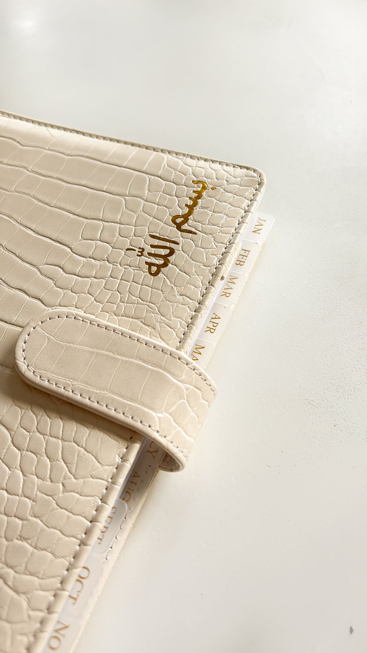 A close-up of a cream-colored crocodile-embossed planner cover with gold foil calligraphy on the front, secured by a matching strap, resting on a light background.