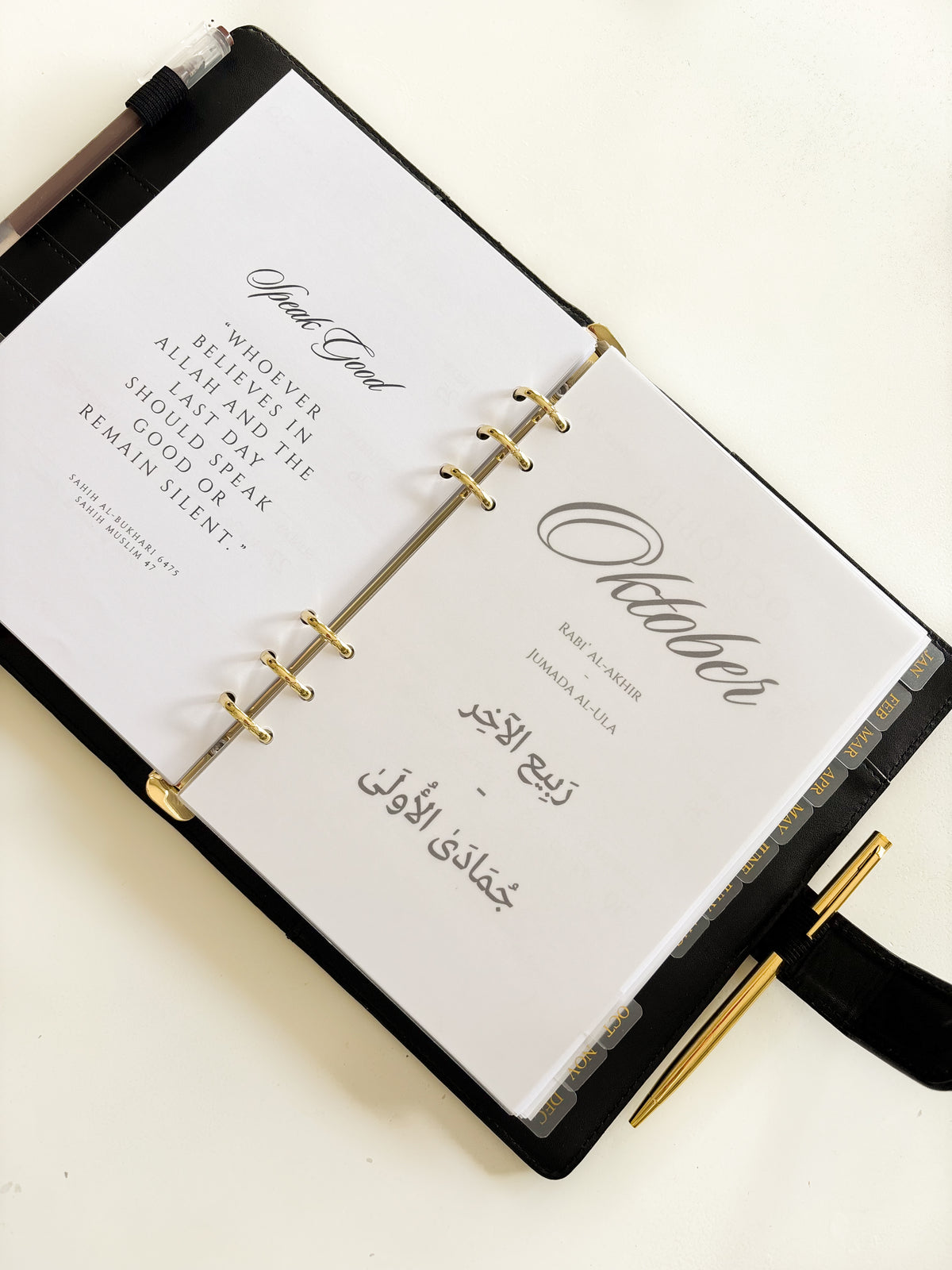 An open black ring-bound planner or journal on a light cream background, showing white interior pages with elegant black script and quote text. Gold-tone rings and pen loops are visible, along with a black pen attached on the right side.