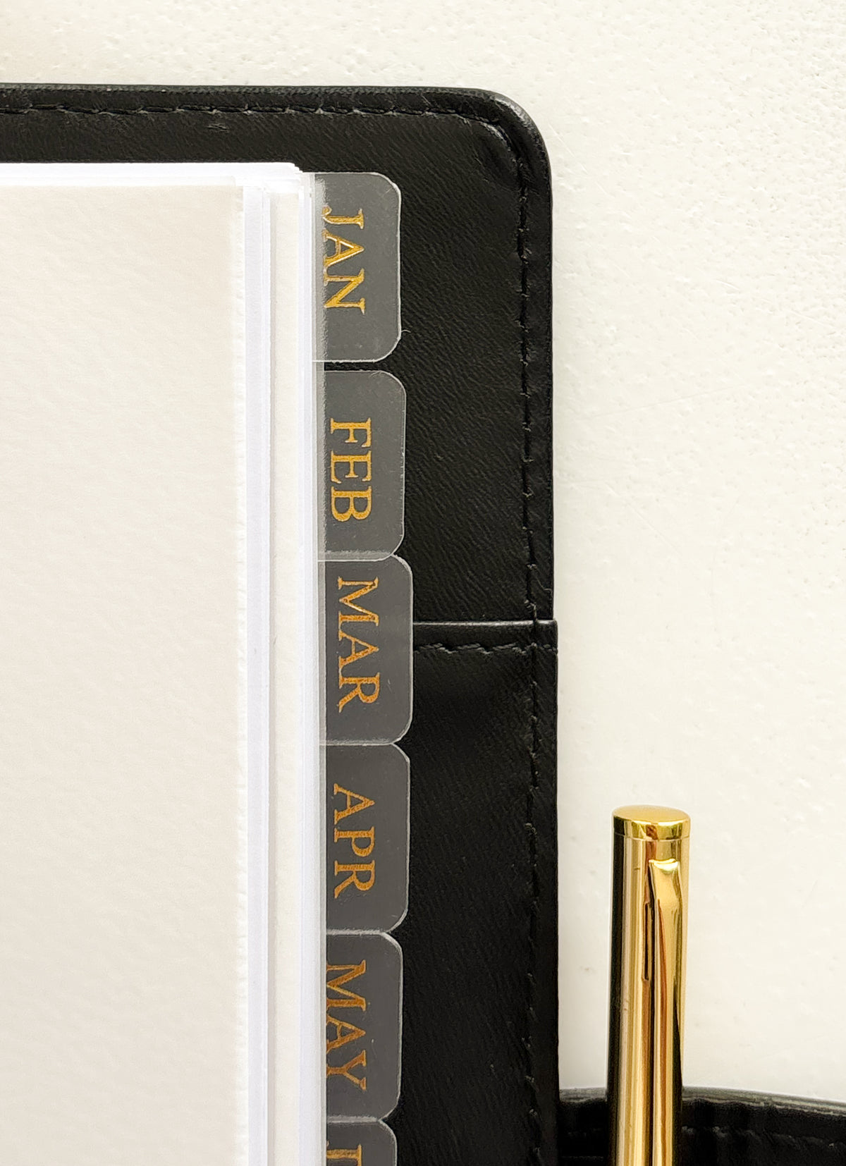 Close-up of a black leather planner or organizer with monthly tab dividers labeled JAN through MAY in gold lettering, next to a gold pen on a light neutral background.