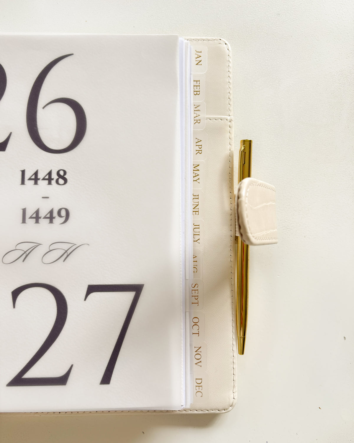 A close-up of a white leather journal or planner cover with large black numbers on the front, a vertical spine printed with months in gold, and a gold pen tucked into a side loop with a white clip.