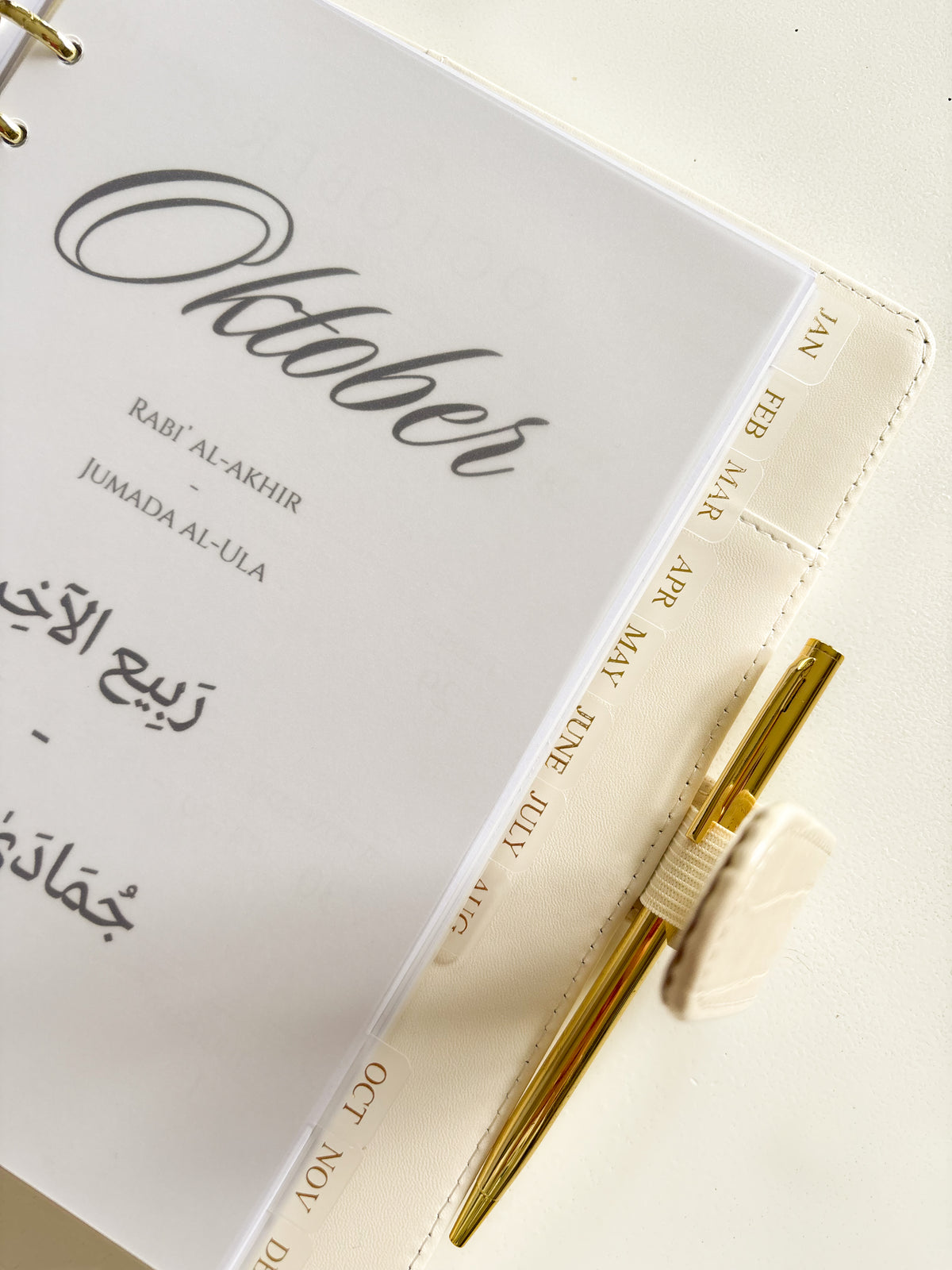 A close-up of a cream-colored monthly planner or journal page with gold lettering, featuring the month 'October' and Arabic text, with a gold pen resting on the right side.